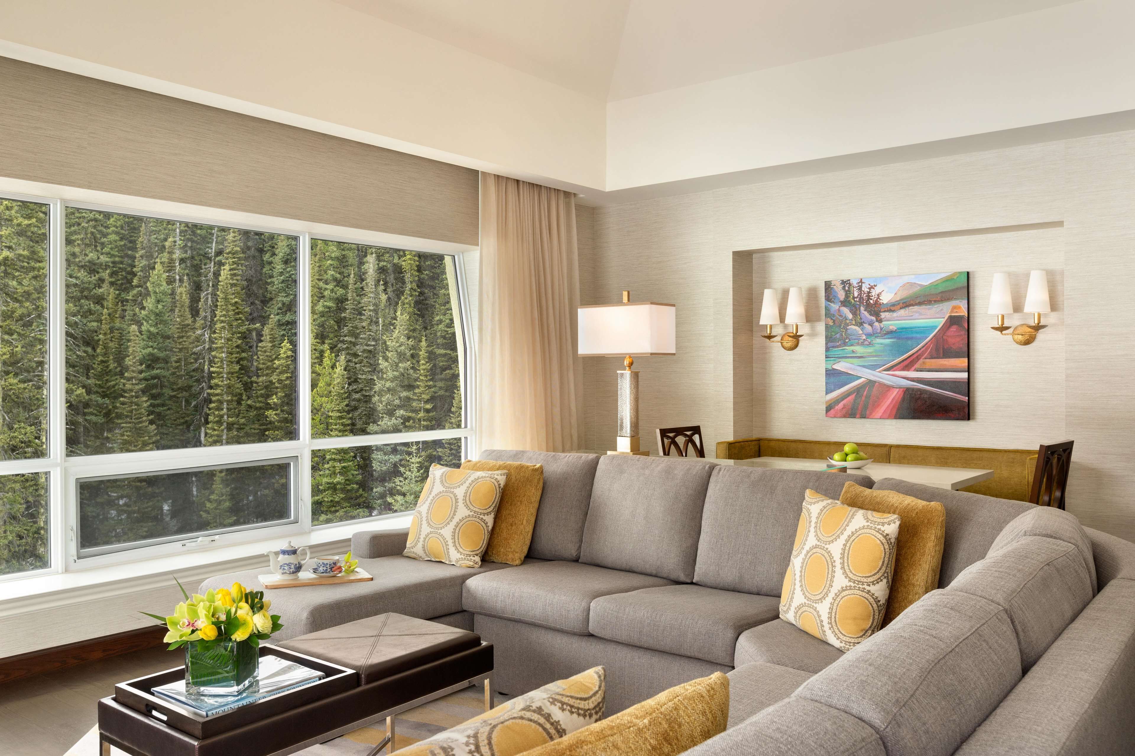 Ein modernes Wohnzimmer mit großem Fenster, einem grauen Ecksofa, einem Beistelltisch mit Blumen und einem Gemälde an der Wand.