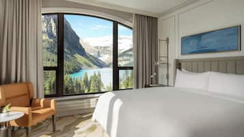 A hotel room with a large bed, a chair, a small table, and a view of mountains and a lake.