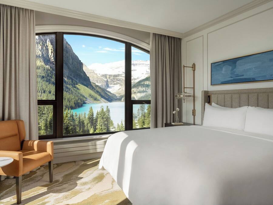 A hotel room with a large bed, a chair, a small table, and a view of mountains and a lake.