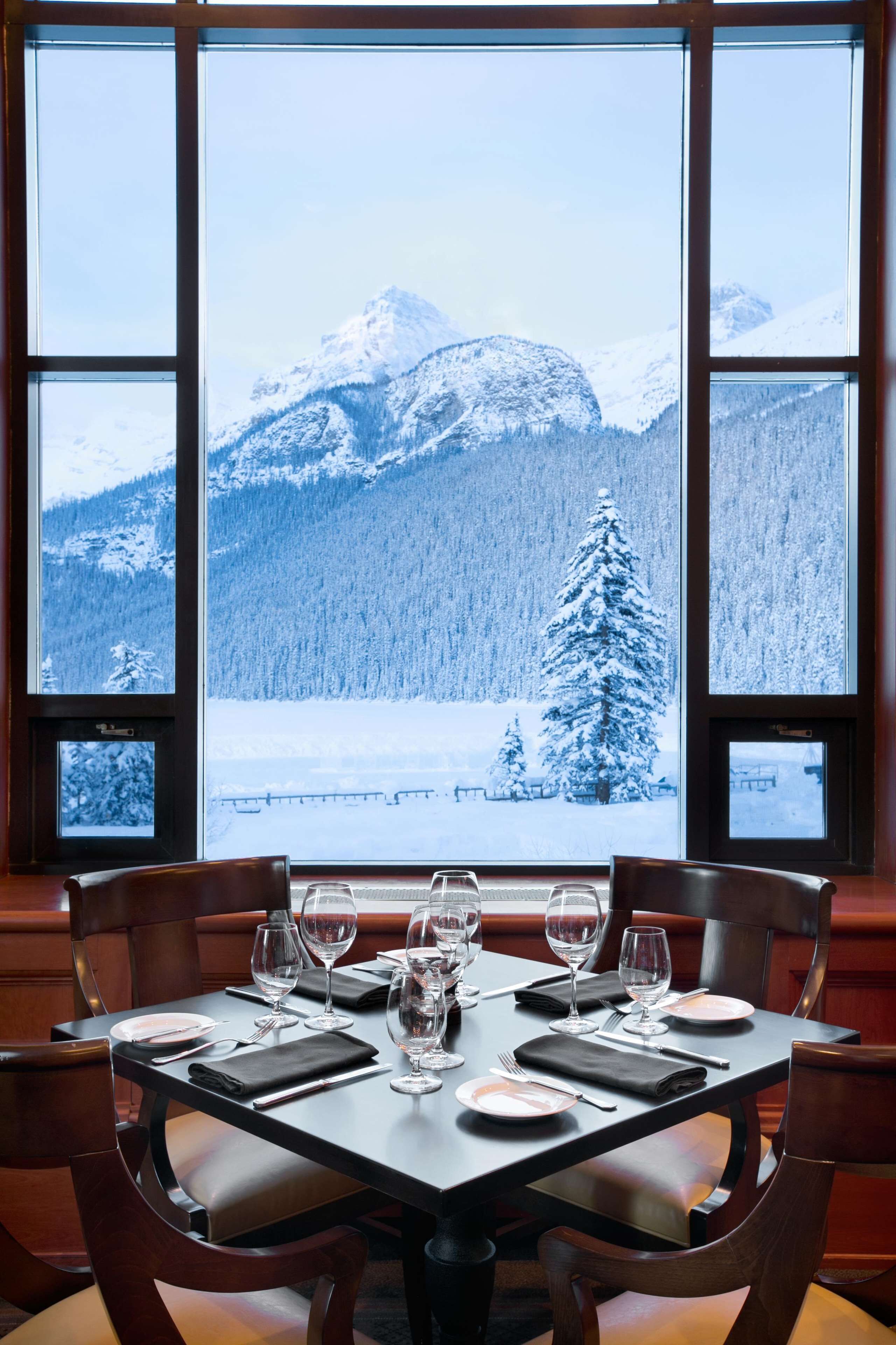 A dining table set for six with a view of snow-covered mountains and a pine tree through large windows.