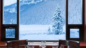 A dining table set for six with a view of snow-covered mountains and a pine tree through large windows.