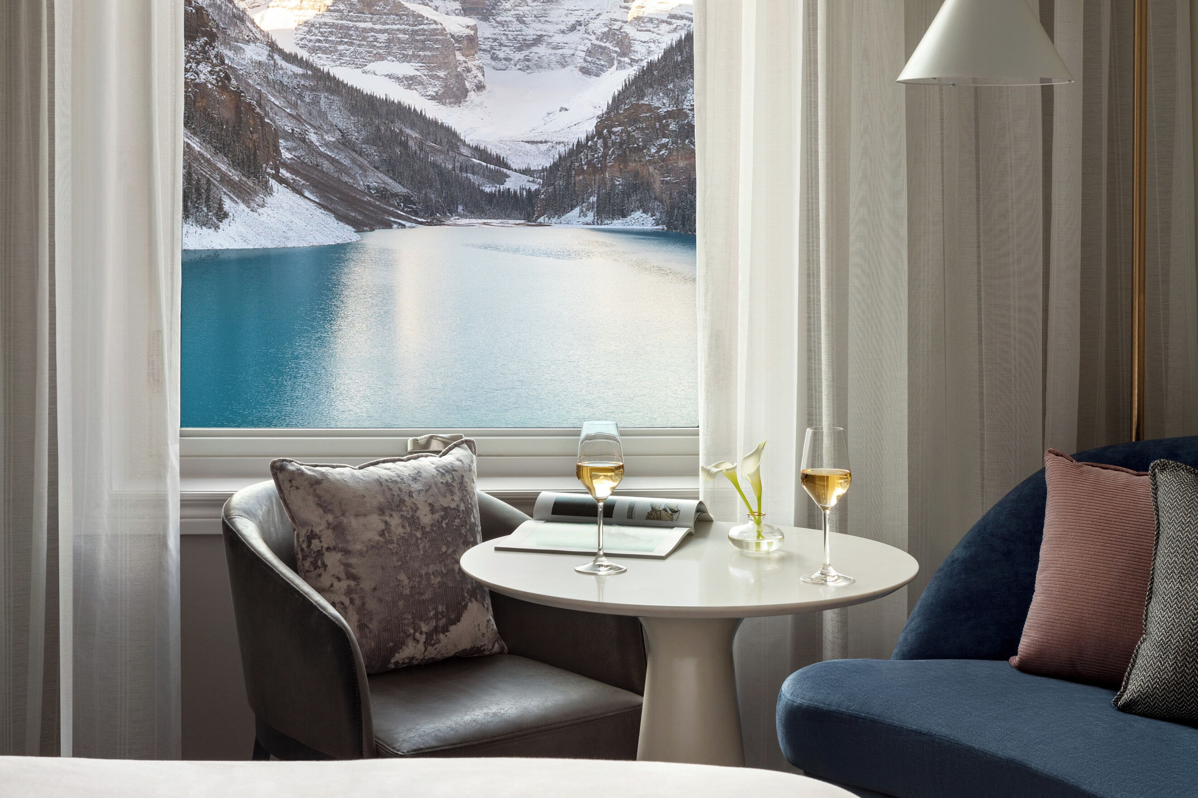 A room with a view of a lake and mountains, a sofa, a chair, and a table with glasses of wine.