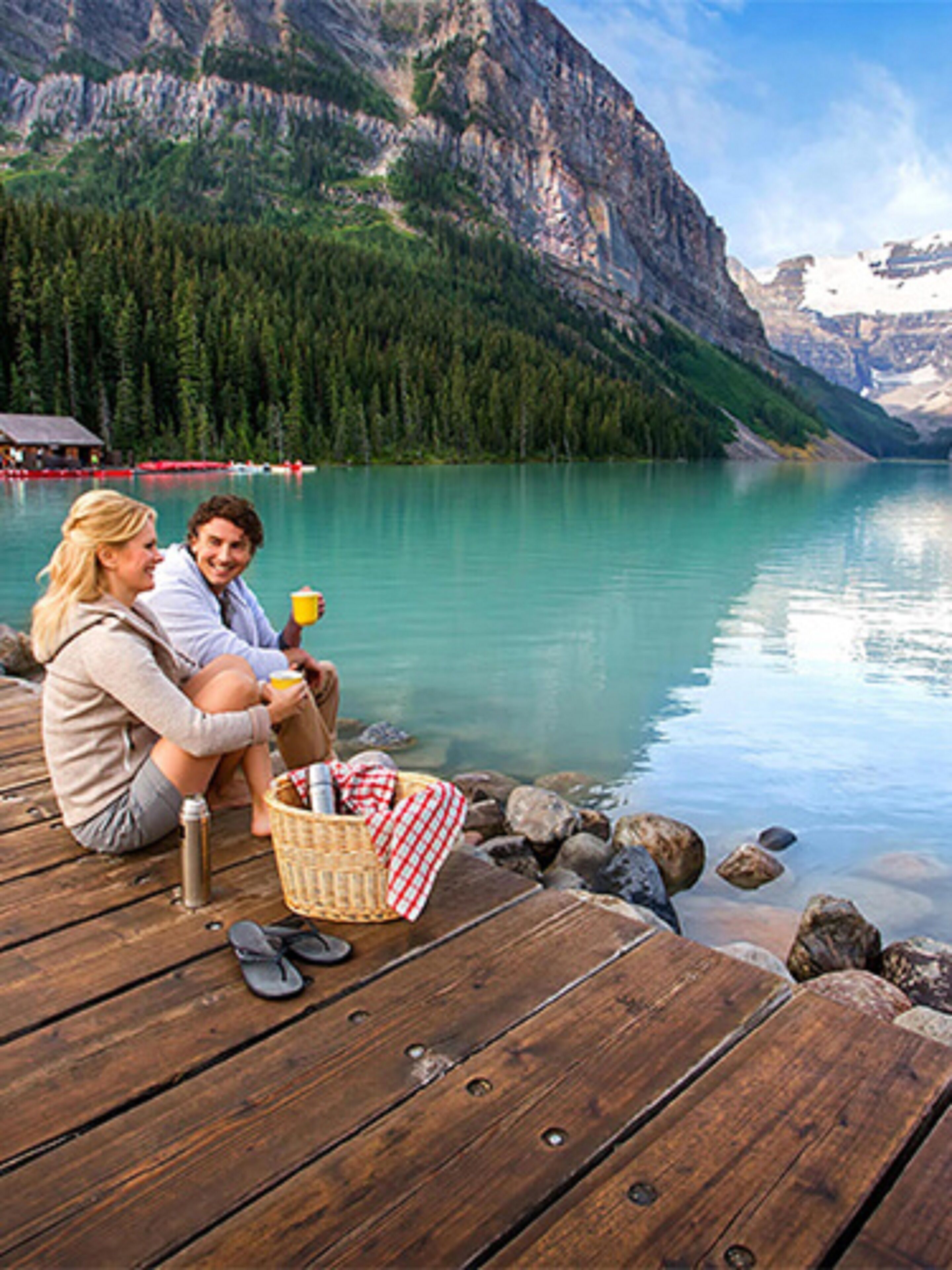 Una pareja sentada en un muelle de madera junto a un lago, con montañas al fondo.