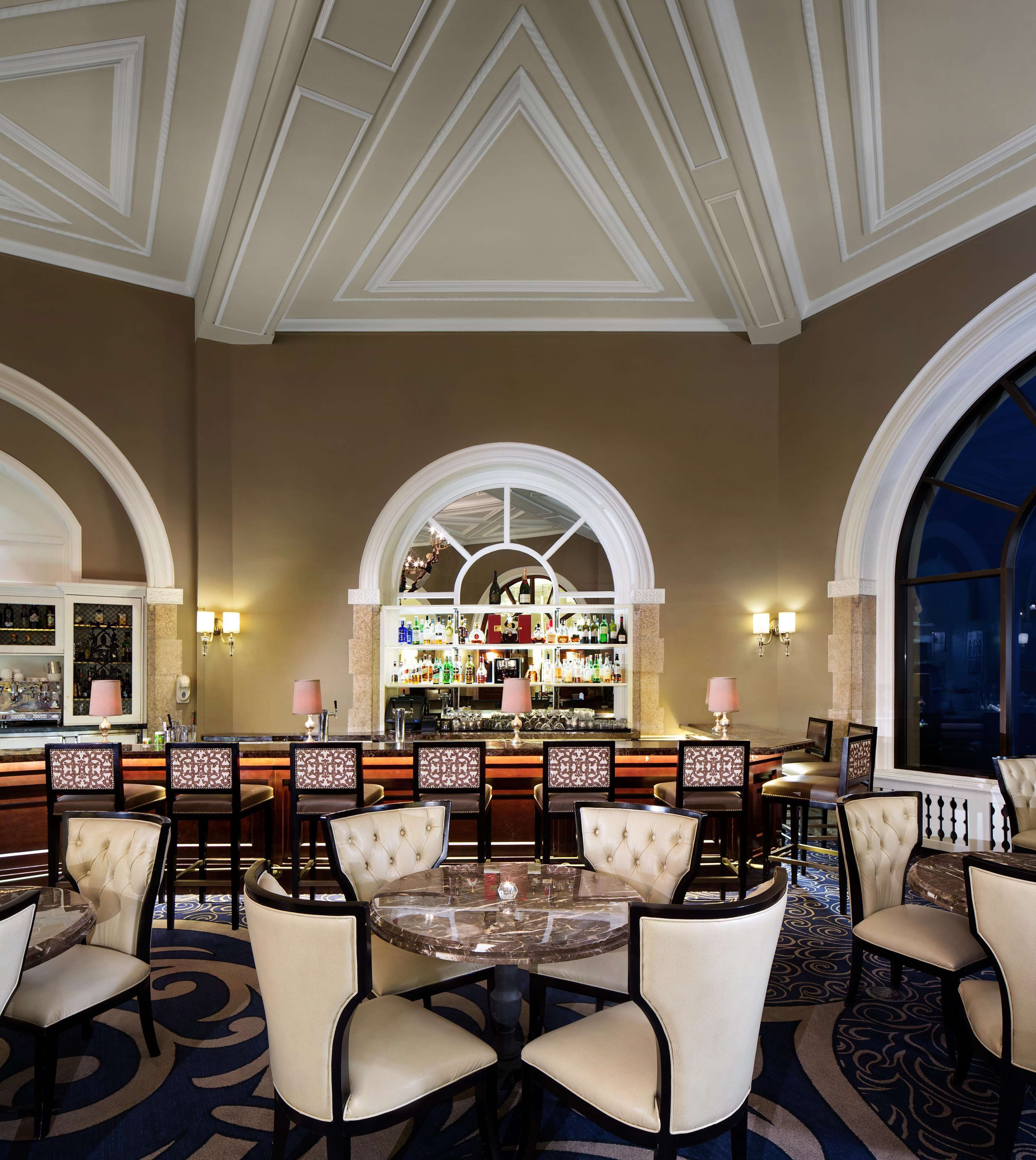 A bar area with a round table and chairs, a bar counter with a wine and liquor display, and arched windows.