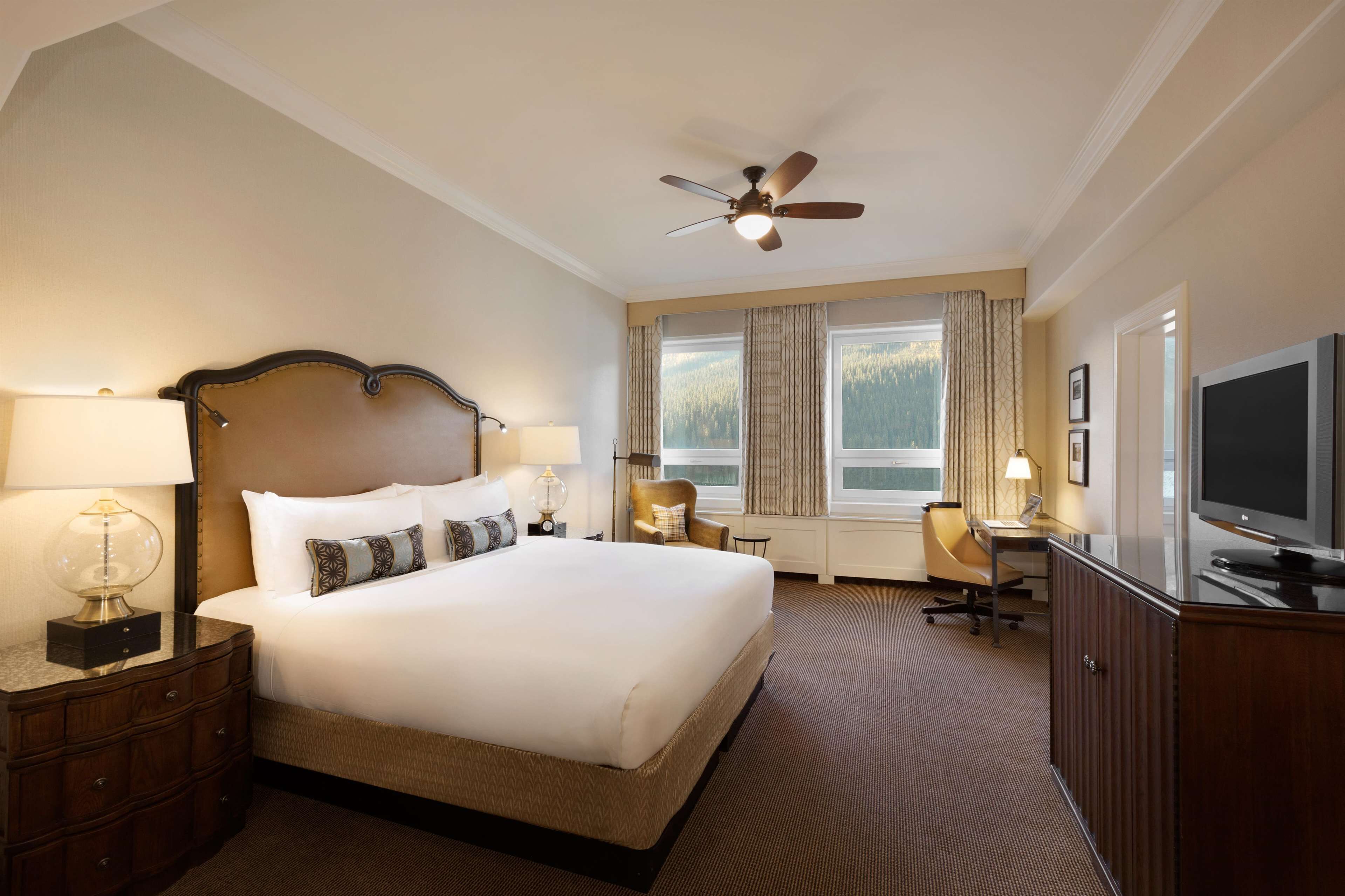 A hotel room with a large bed, a desk with a chair, a television, and a ceiling fan.