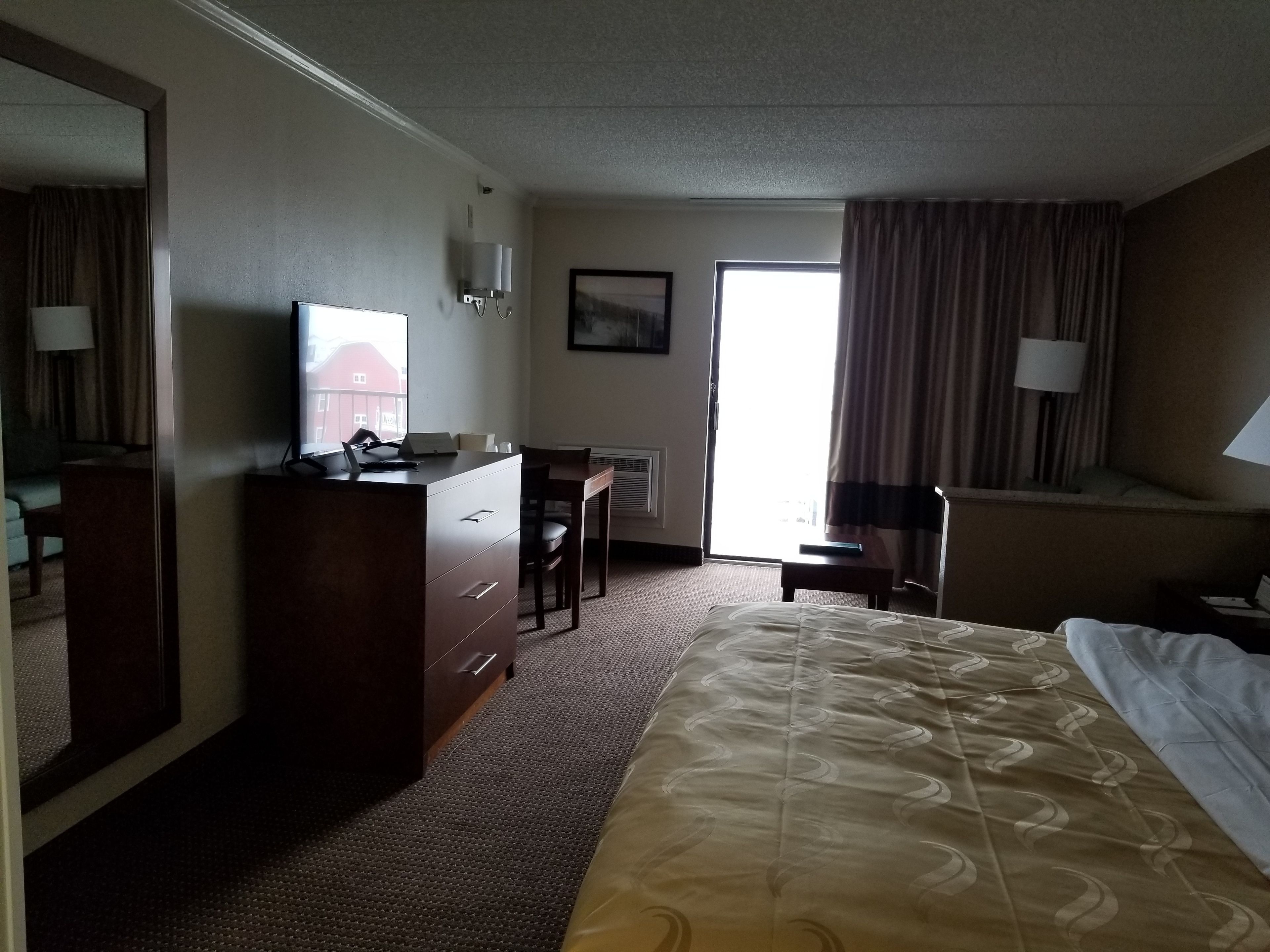 A hotel room with a bed, a desk, a chair, a dresser, a mirror, a window with curtains, and a wall-mounted lamp.