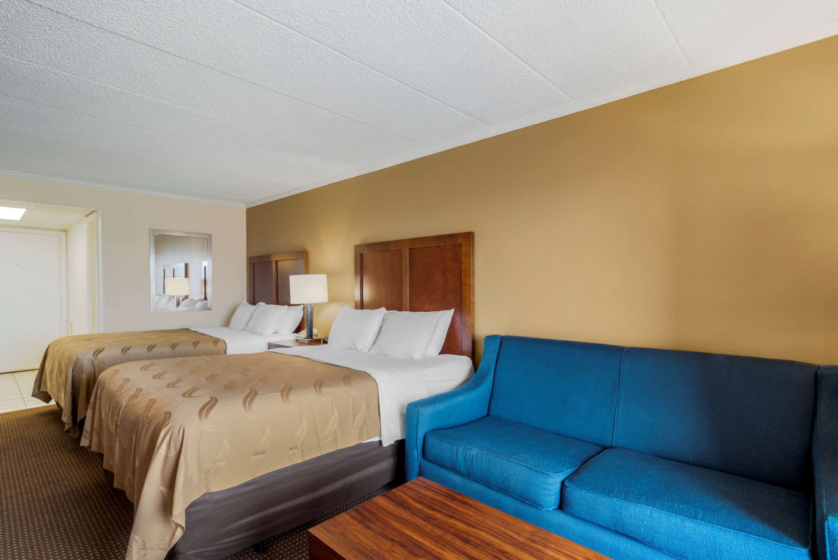Quarto de hotel com duas camas, um sofá azul, uma mesa de centro de madeira e uma mesinha de cabeceira com abajur.