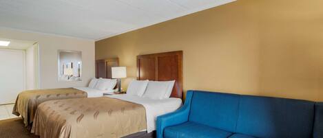Quarto de hotel com duas camas, um sofá azul, uma mesa de centro de madeira e uma mesinha de cabeceira com abajur.