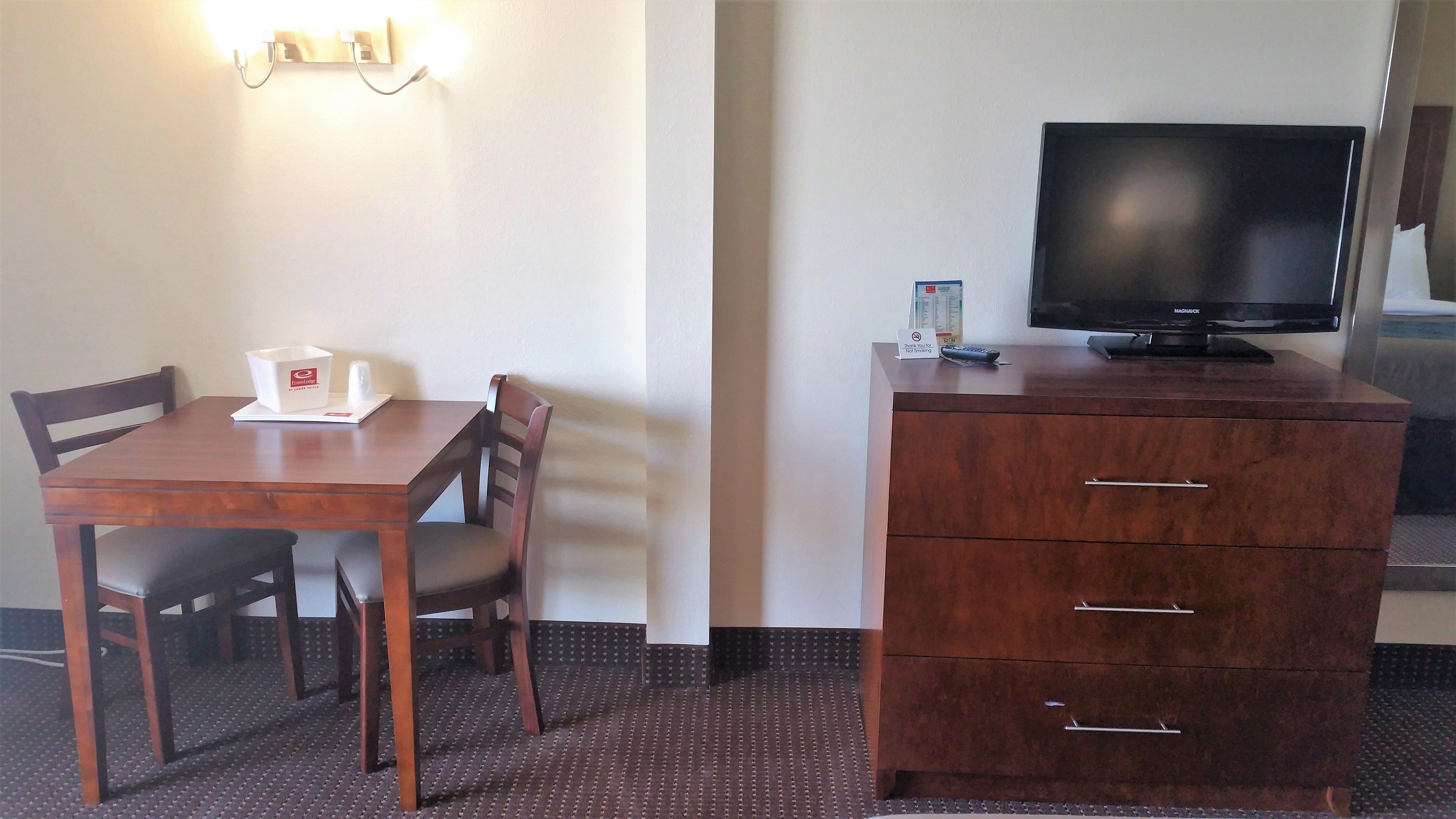 A hotel room with a wooden desk, two chairs, a television on a dresser, and a sign indicating no smoking.