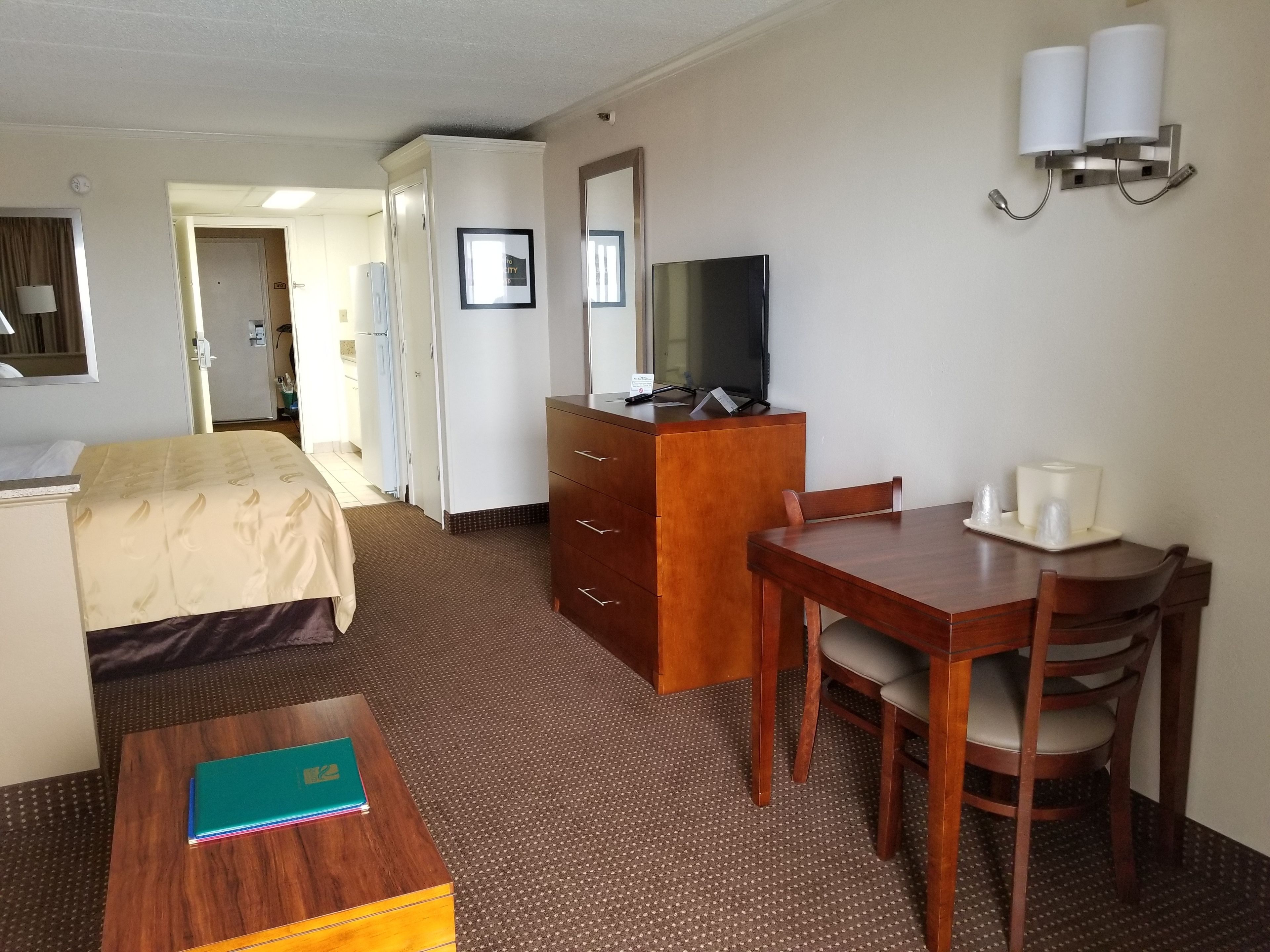 A hotel room with a bed, a desk with a chair, a dresser, a television, and a wall-mounted lamp.