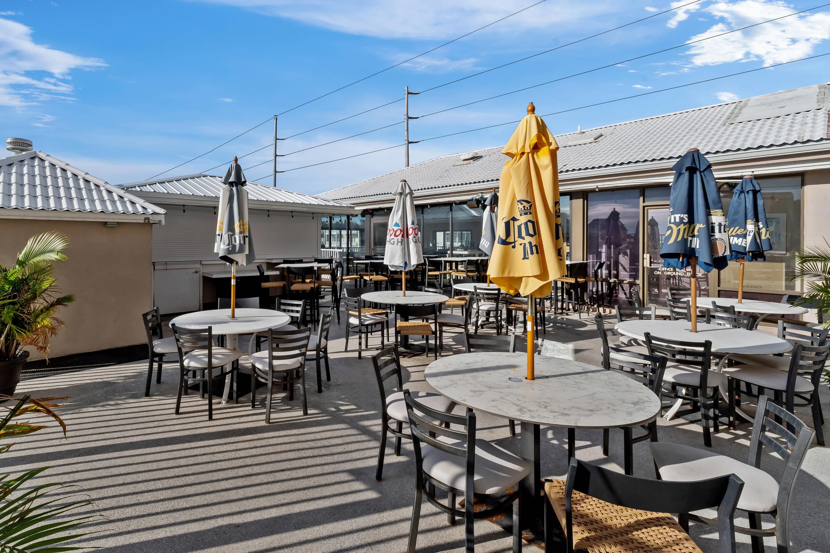 An outdoor café area with round tables and chairs, umbrellas, and a building with a tiled roof in the background.