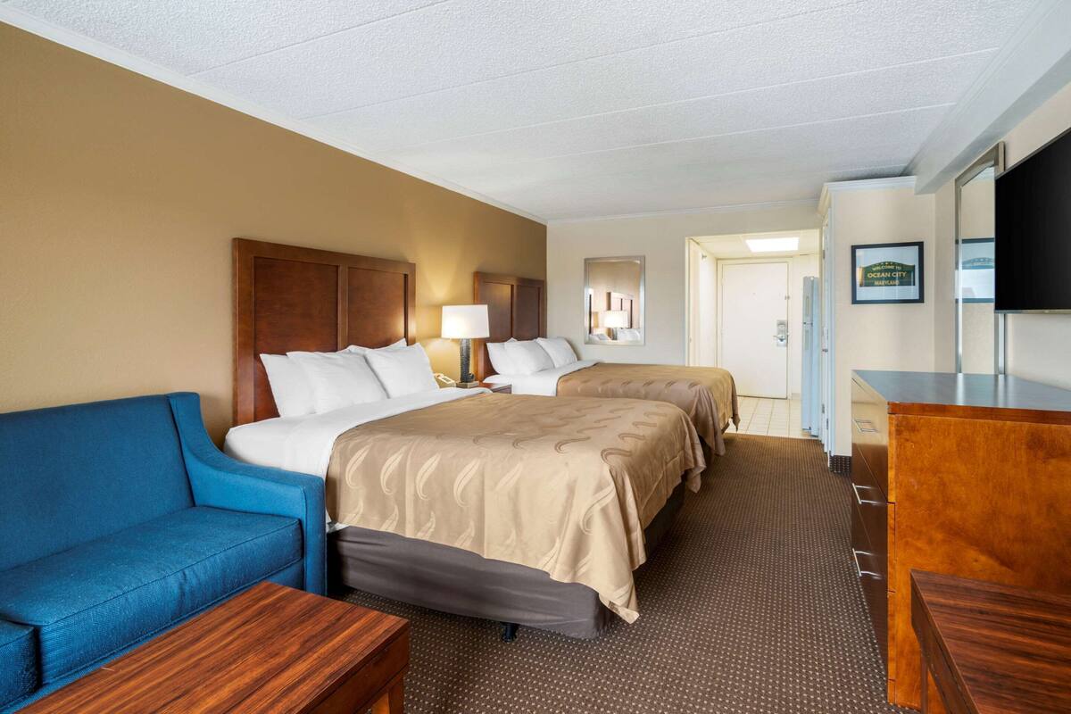 A hotel room with a blue sofa, two beds with beige bedding, a wooden desk, a mirror, and a television.