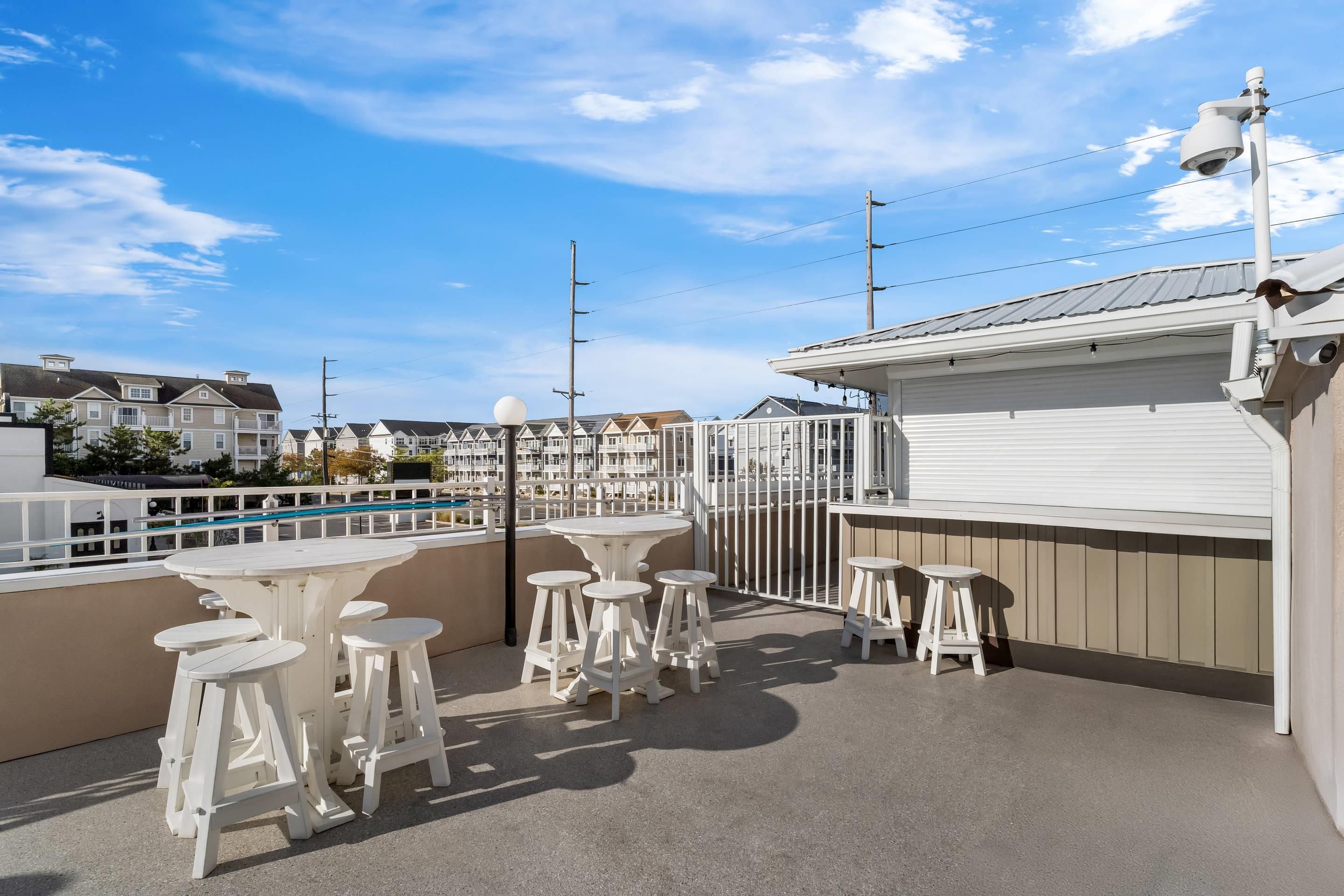 Eine Dachterrasse mit weißen Tischen und Hockern, im Hintergrund ein Pool und Blick auf Wohnhäuser.