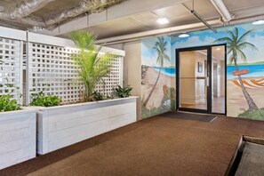 Un espace d’accueil agrémenté d’un grand pot contenant un palmier, un mur orné d’une fresque représentant une plage et une porte portant l’inscription « BUREAU ».