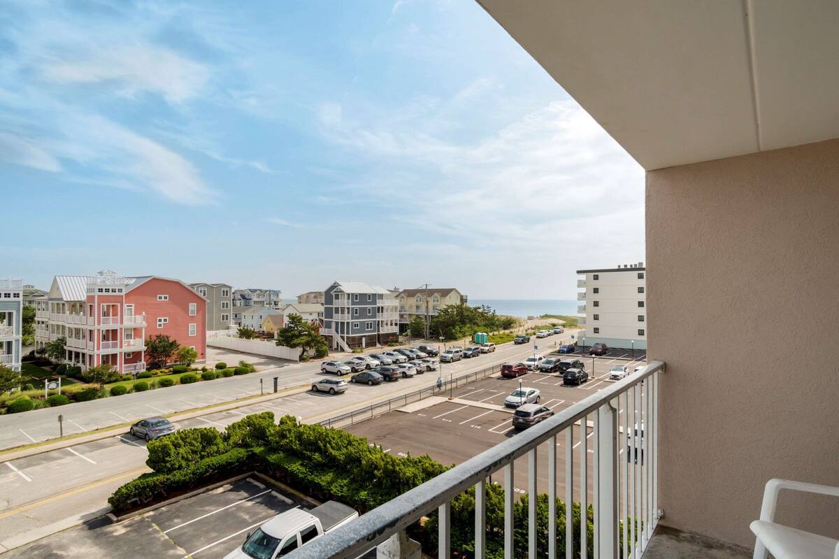 A balcony with a view of a parking lot and buildings, including a red building and a white building with a balcony.