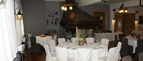 A dining area with round tables set for a meal, white chairs, and a stone fireplace.