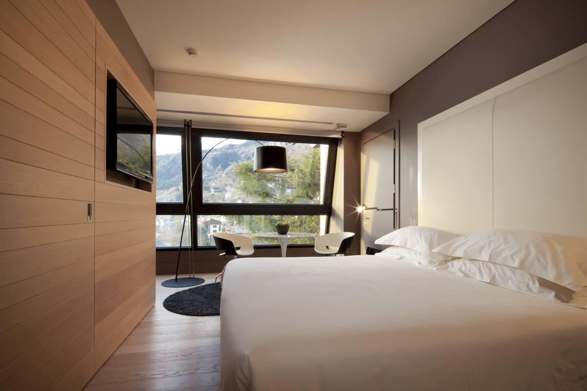 Un dormitorio moderno con una cama grande, un televisor montado en la pared, una mesita con sillas y vistas a montañas a través de la ventana.