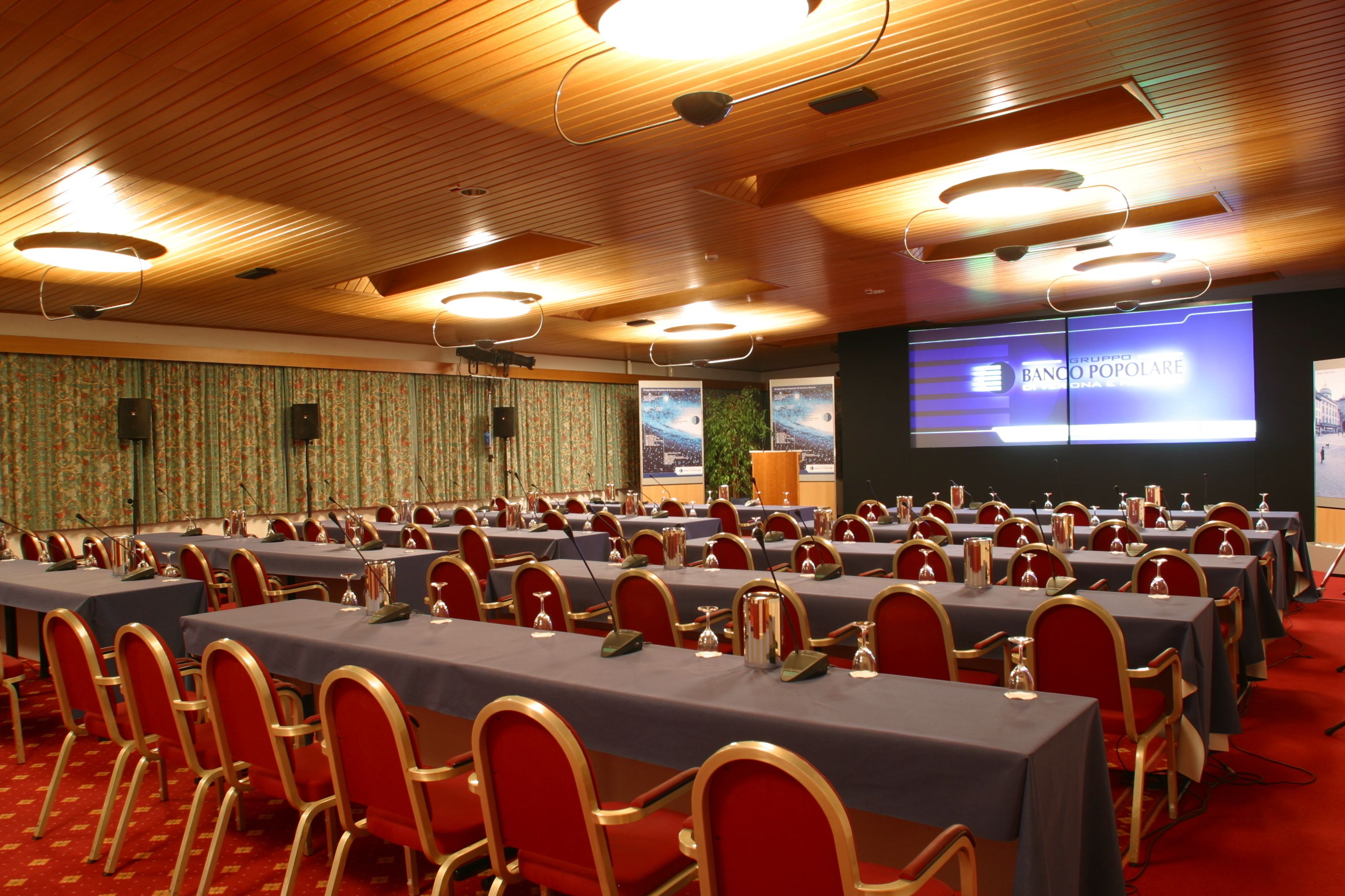 A conference room with multiple tables and chairs, a presentation screen, and a wooden ceiling.