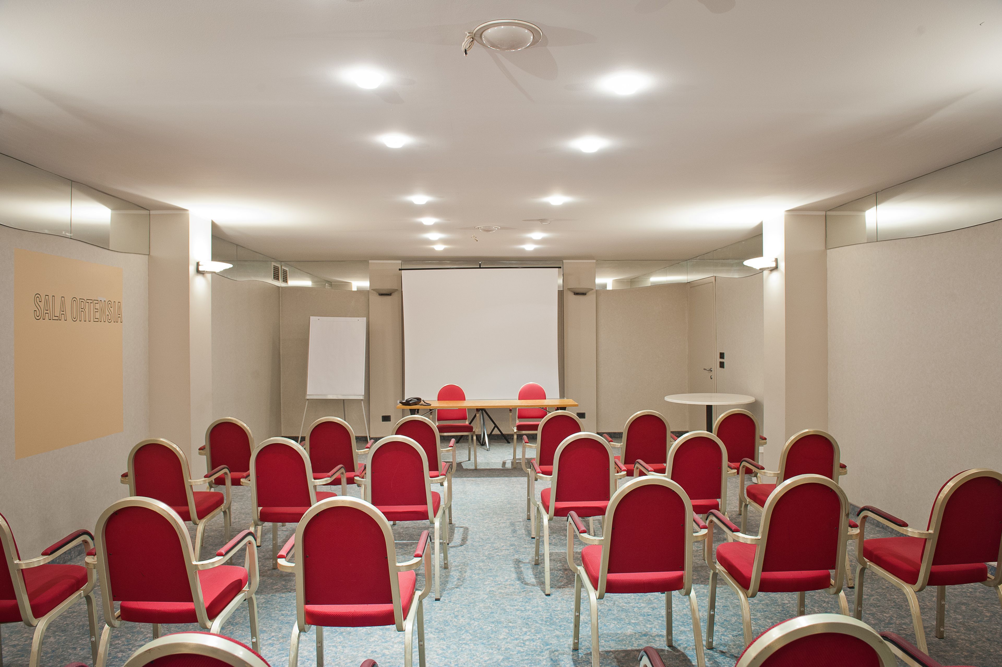 Un salón de conferencias con sillas rojas, una pizarra y una pantalla de proyector.