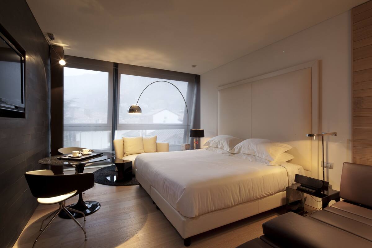 Una habitación de hotel moderna con una cama grande, un escritorio con una silla, una mesita y una ventana con vistas a la ciudad.