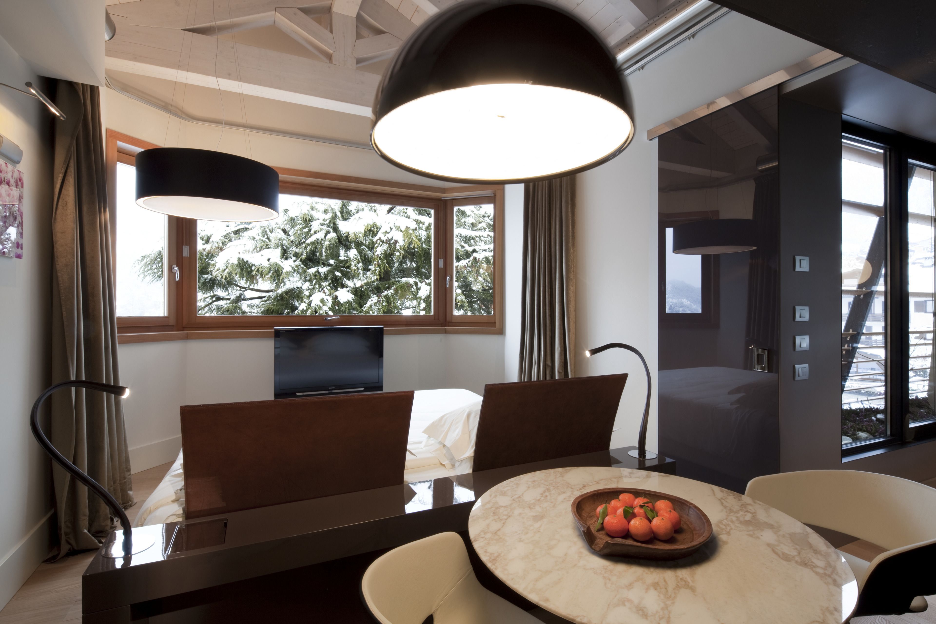A modern hotel room with a dining area, a large window with a view of trees, a flat-screen TV, and a round table with a bowl of fruit.