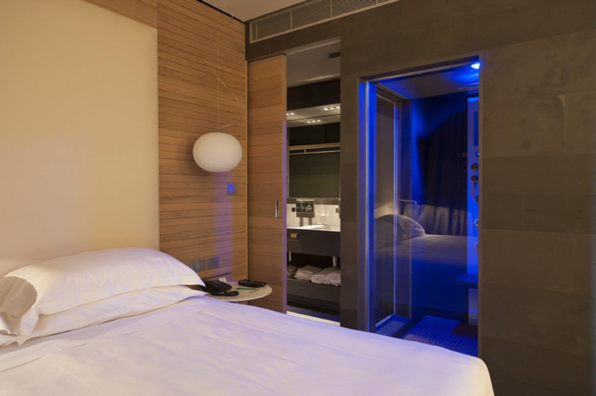 A modern hotel room with a bed, a bedside table, a wardrobe, and a bathroom visible through an open door.