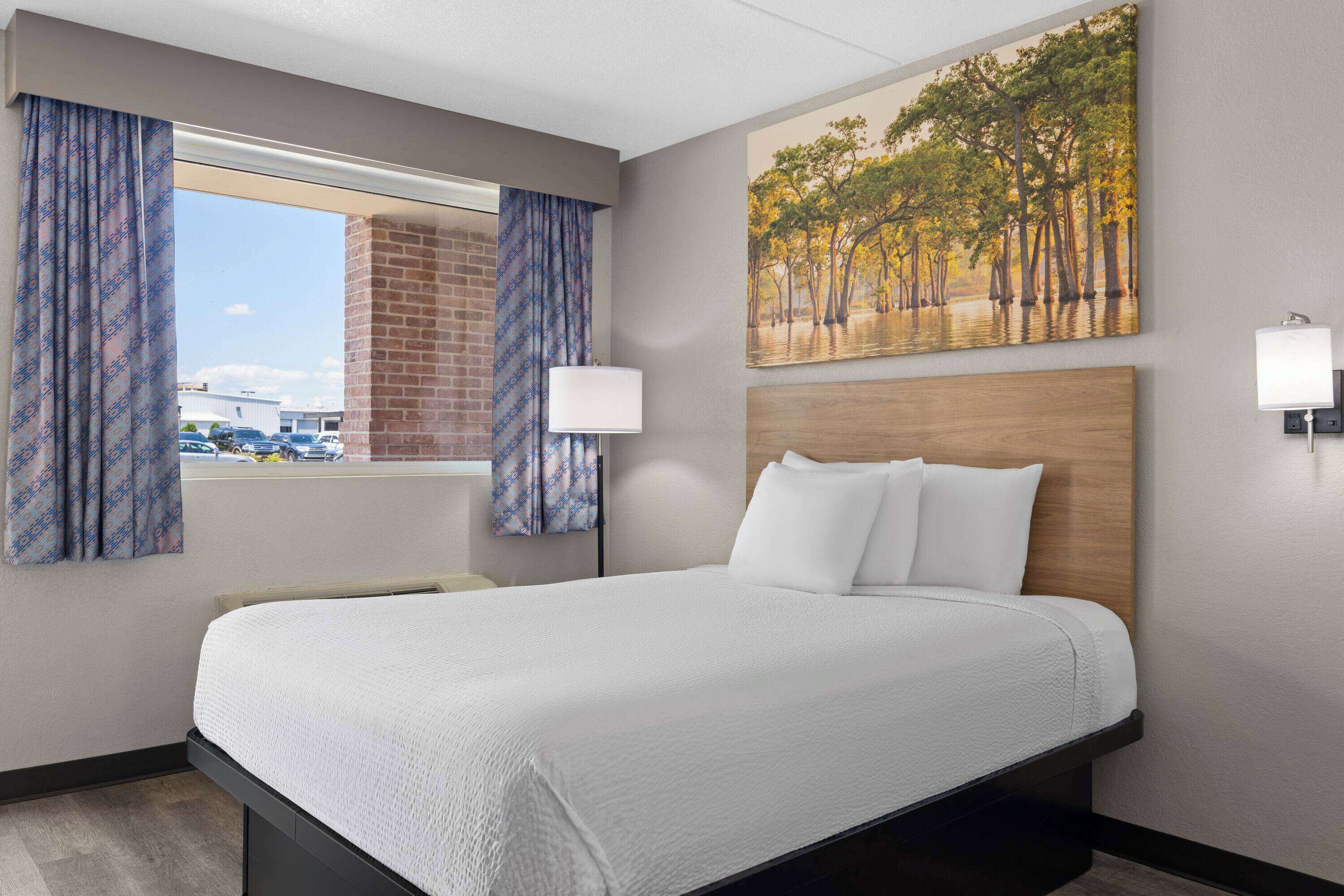 A hotel room with a large bed, a window with curtains, a painting of trees, and a lamp on the wall.