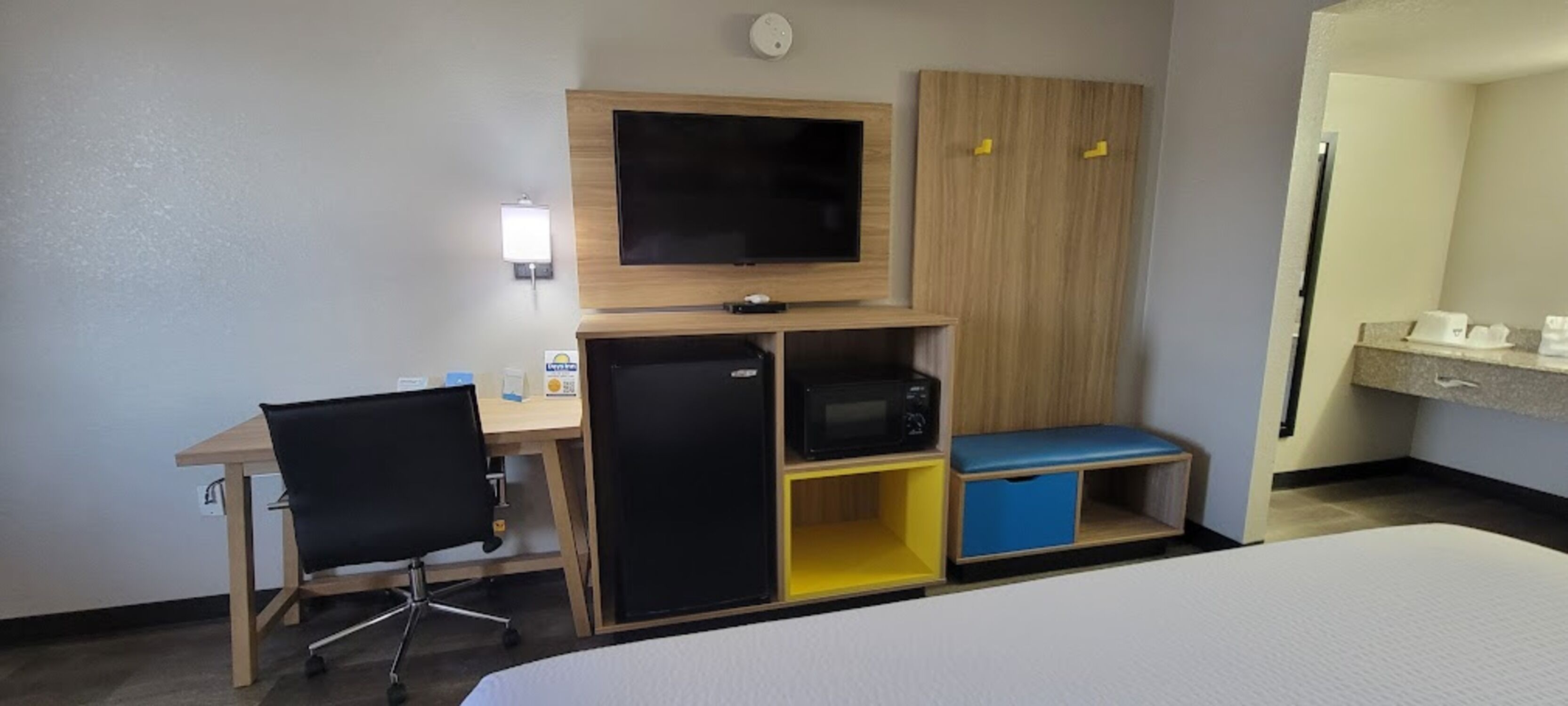 Habitación de hotel con cama, escritorio con silla, televisor, microondas y una pequeña zona de estar con un banco azul.