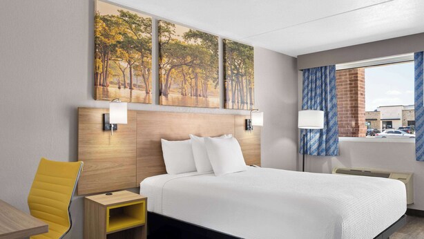 A hotel room with a large bed, a wooden nightstand, a yellow chair, and a window with a view of the street. - Days Inn by Wyndham Conway (Conway)
