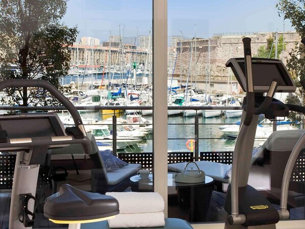 Ein Fitnessraum mit Trainingsgeräten und Blick auf einen Yachthafen mit Booten.