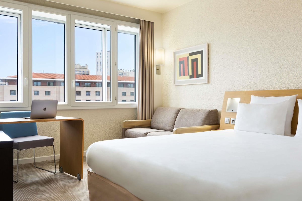 Ett hotellrum med en säng, ett skrivbord med en bärbar dator, en stol och ett fönster med utsikt över staden.