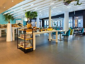Ein modernes Café mit weißen Tischen, smaragdgrünen Stühlen und einer Auswahl an Backwaren auf Regalen.