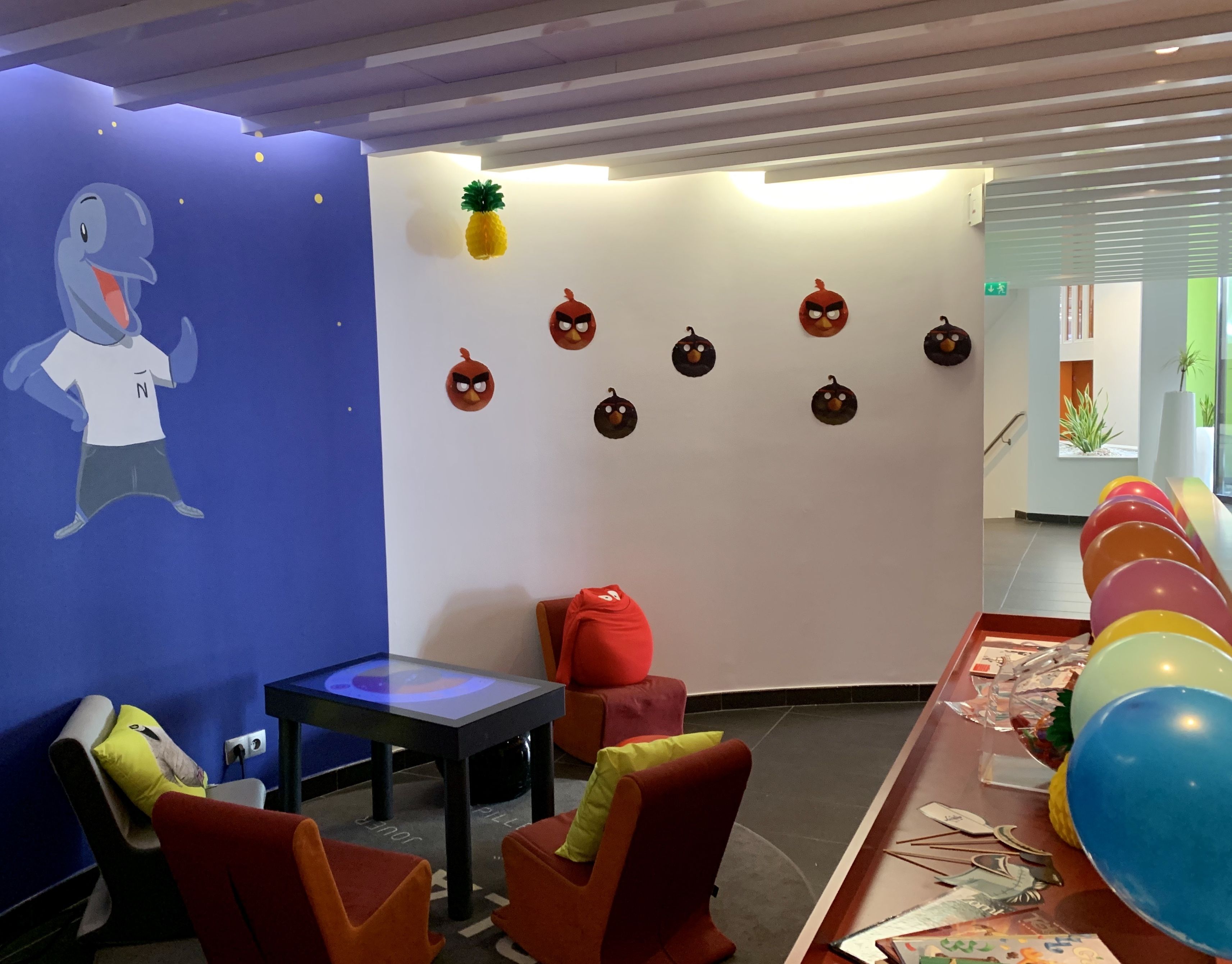 A room with a blue wall, a white wall with Angry Birds decorations, a table with balloons, and a chair with a yellow pillow.