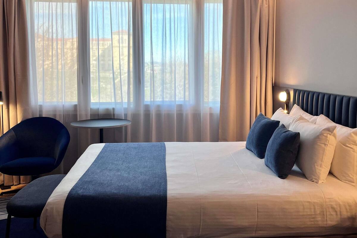 Habitación de hotel con cama, una silla, una mesita y vistas a través de cortinas translúcidas.