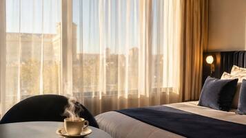 A hotel room with a bed, a round table with a cup of coffee, and a view of the city through large windows.
