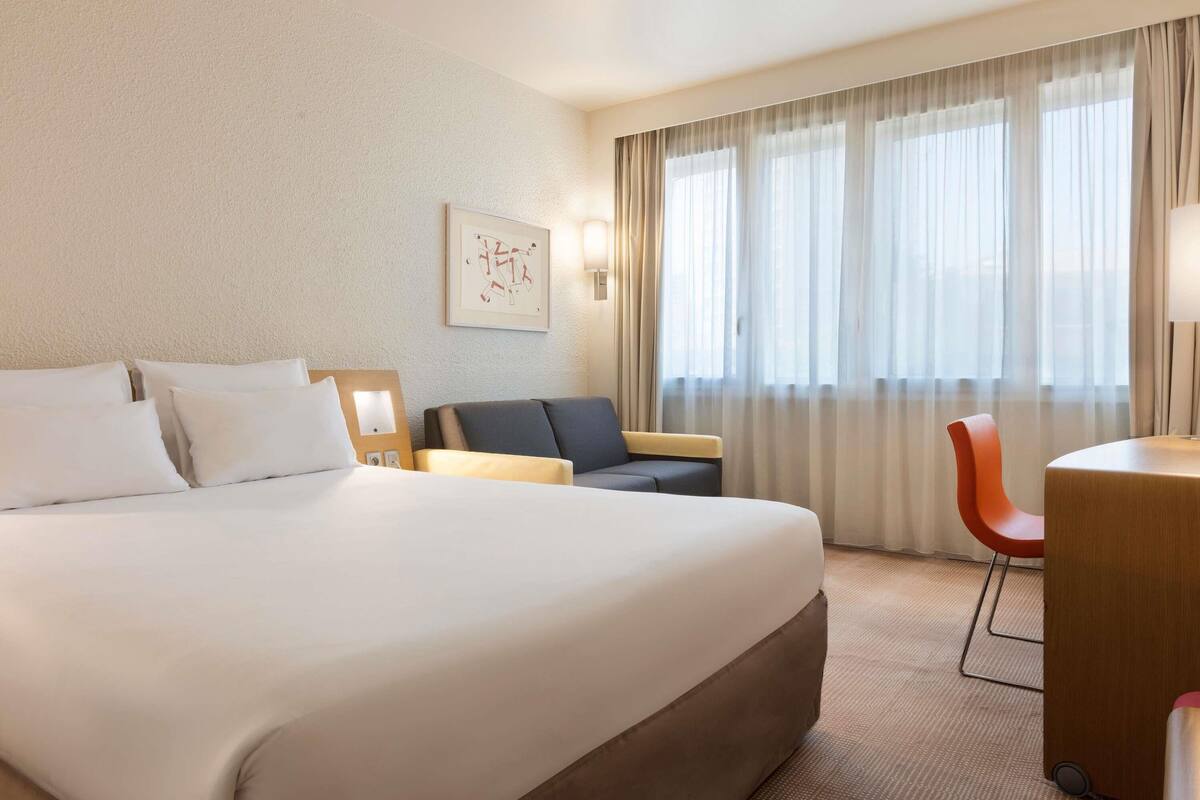 Une chambre d’hôtel avec un grand lit, un bureau, deux chaises et une fenêtre avec des rideaux.