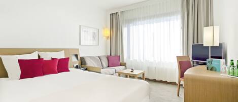 A hotel room with a large bed, a sofa, a small table, a TV, and a desk with a lamp.