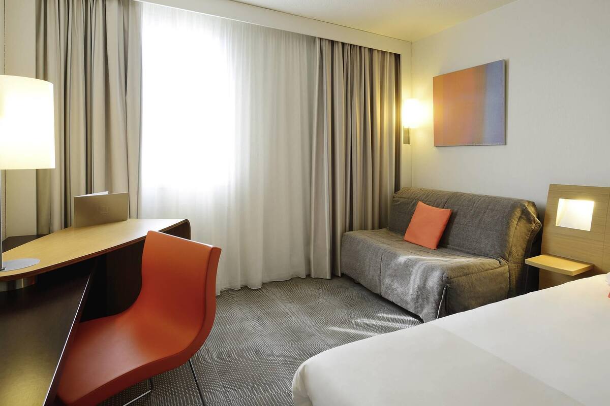 Habitación de hotel con cama, escritorio, silla, sofá y un gran ventanal con cortinas.