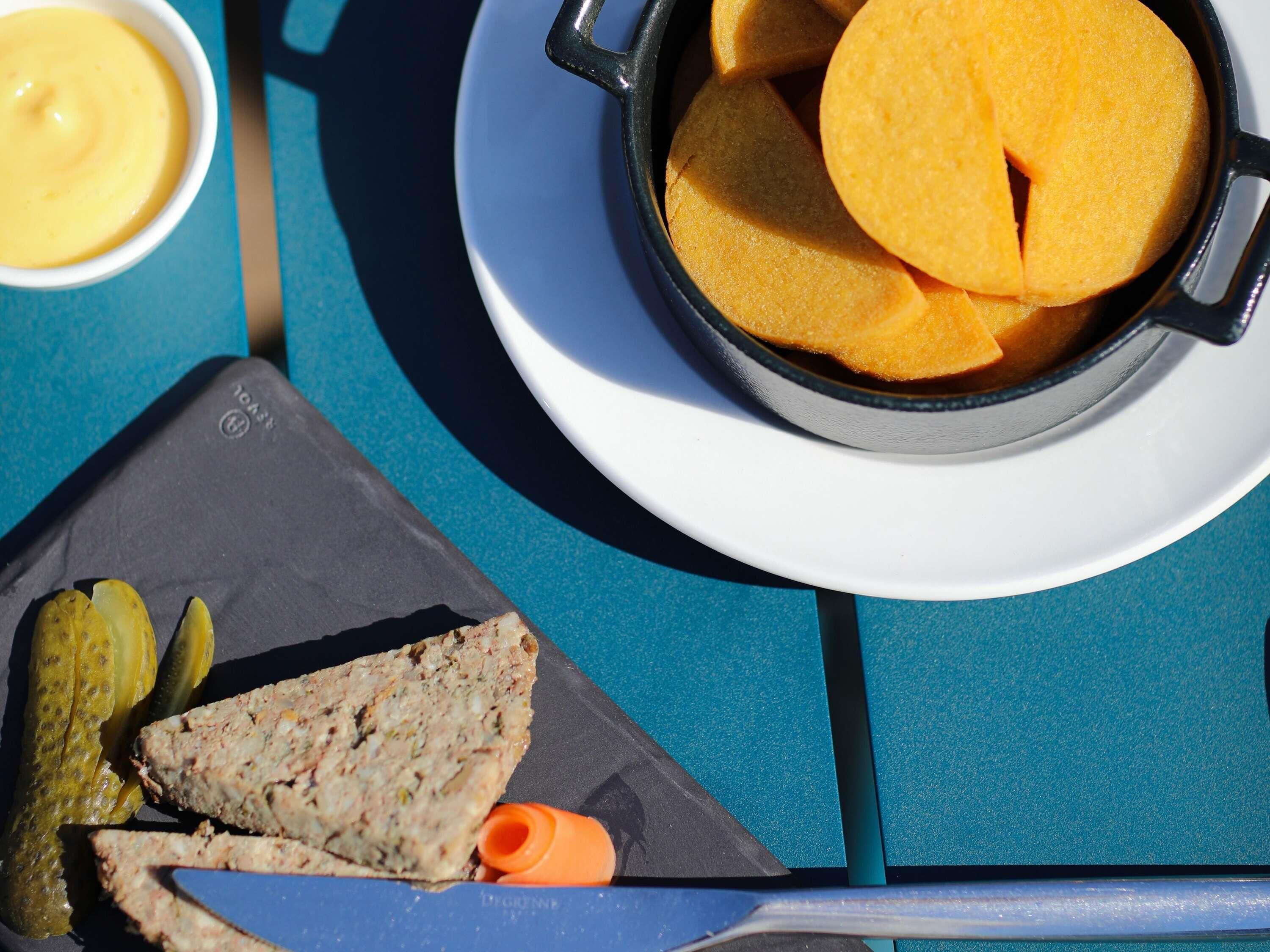 A table with a black pan filled with yellow potato wedges, a white plate with a slice of potato wedge, a small bowl of yellow sauce, and a plate with a meatloaf slice, pickles, and a carrot.