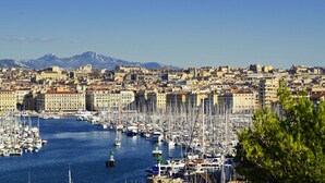 Un port de plaisance rempli de nombreux bateaux, entouré d’un quartier urbain densément peuplé, avec des bâtiments historiques et une montagne en arrière-plan.