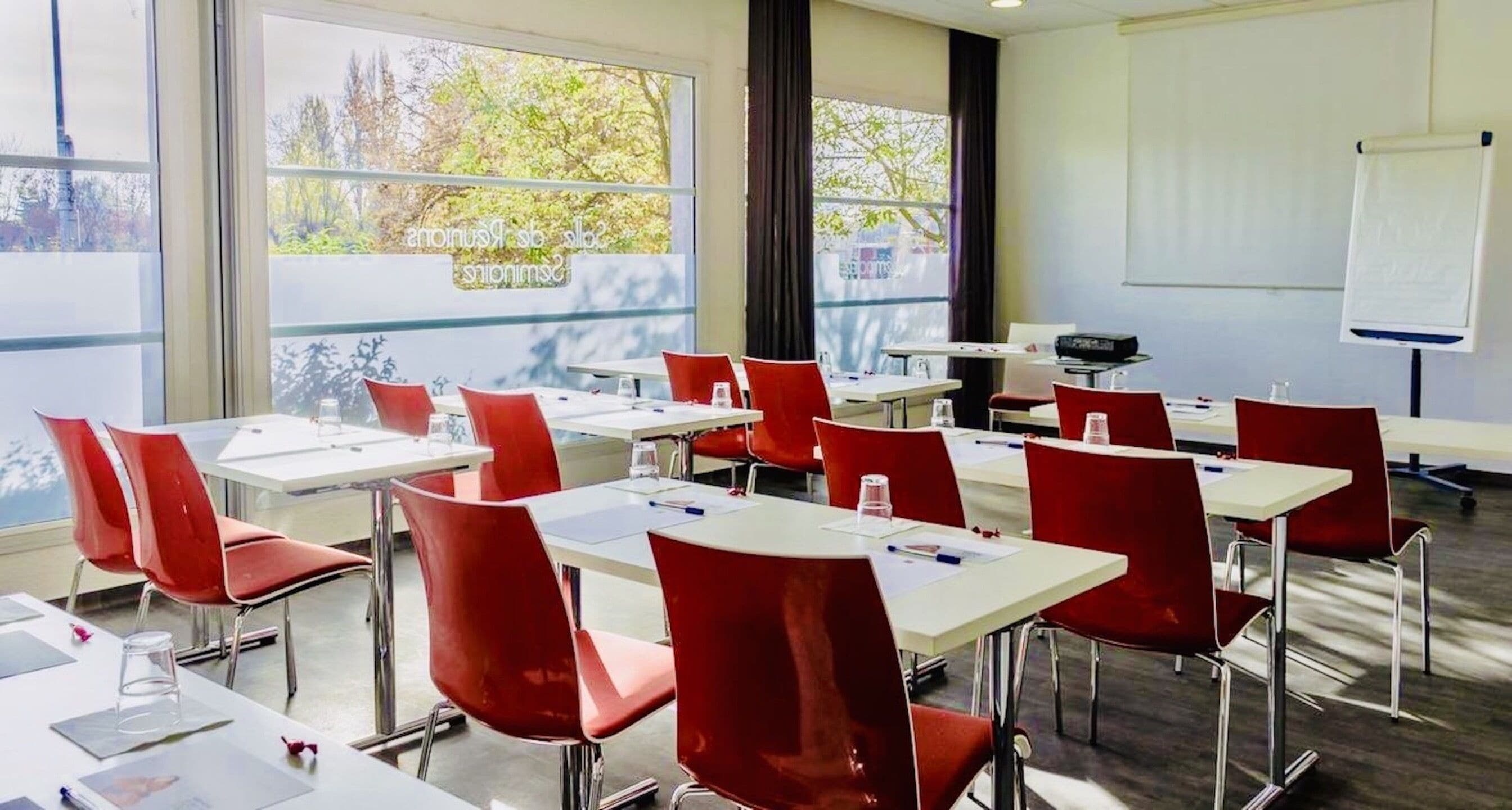 A conference room with white tables and red chairs, large windows with a view of trees, and a whiteboard on the wall.