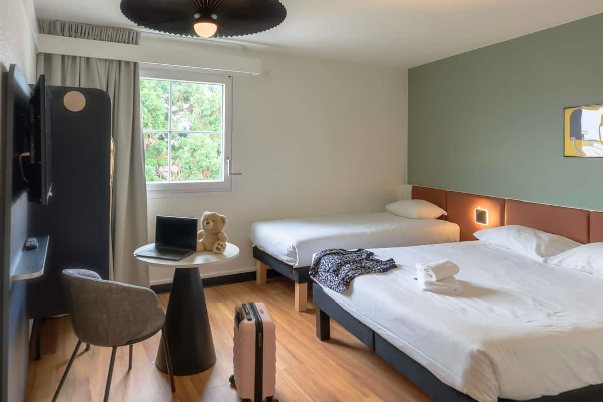 Een hotelkamer met een bed, een bureau met een laptop, een stoel, een koffer en een raam met gordijnen.