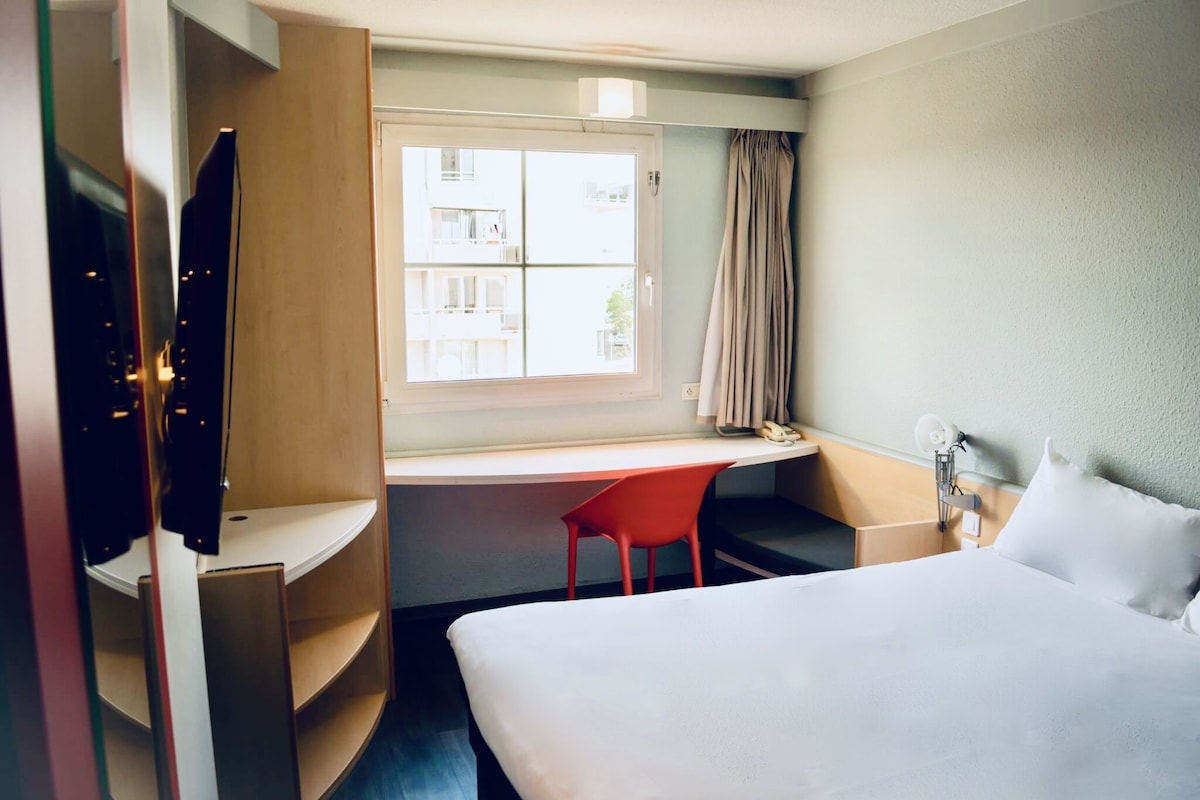 Een hotelkamer met een bed, een bureau, een stoel, een televisie en een raam met gordijnen.