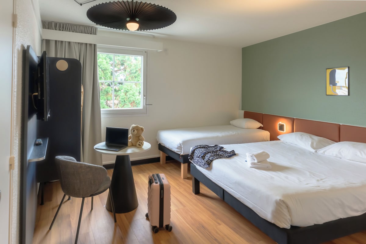Een hotelkamer met twee bedden, een bureau met een laptop, een stoel, een koffer en een knuffelbeer.