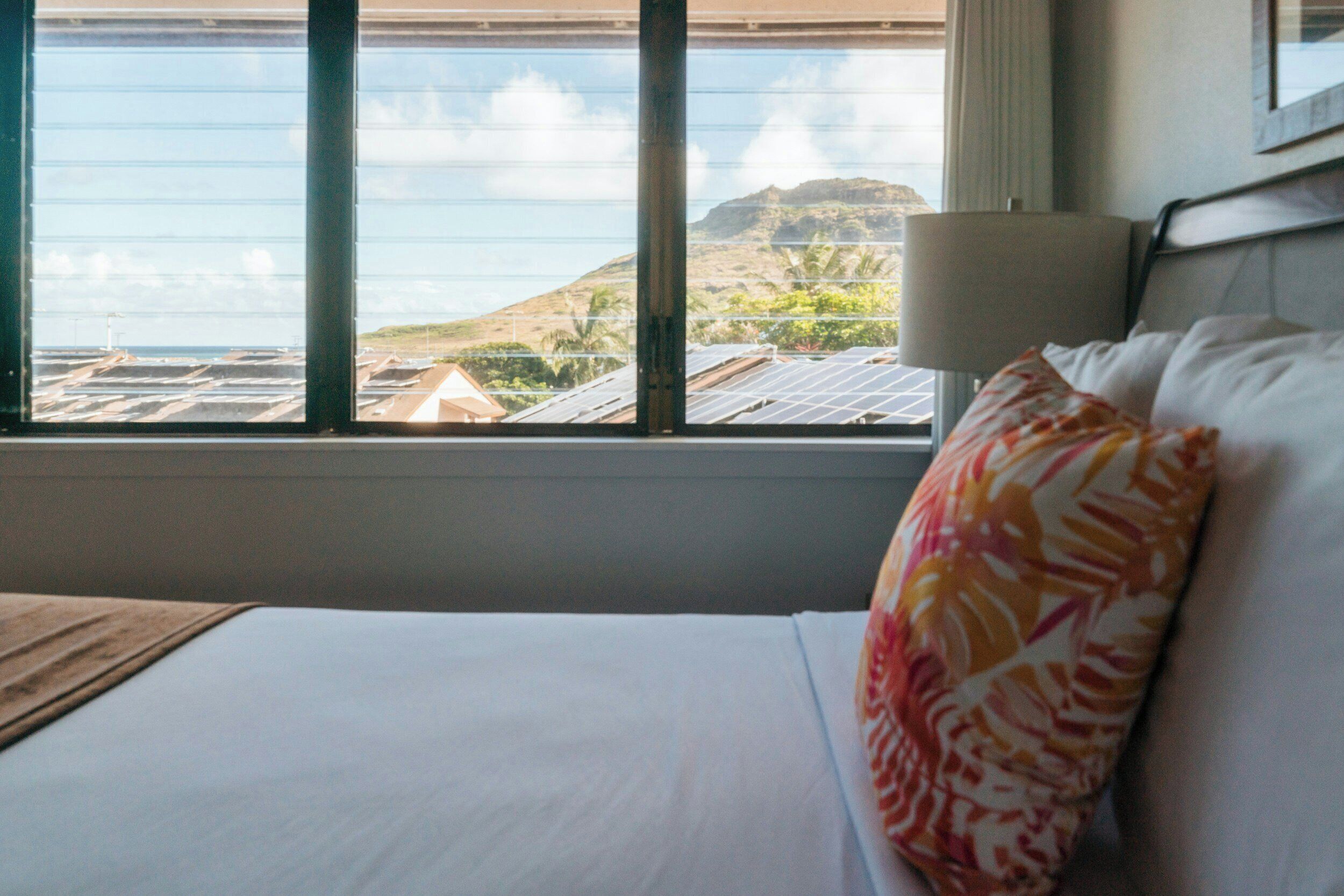 A hotel room with a bed, a window with a view of the ocean and a distant mountain, and a lamp on the bedside table.