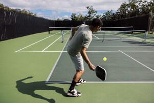 Una cancha de tenis con red, un jugador con camisa y pantalones cortos de color claro, y una pelota de tenis en movimiento.
