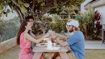 A picnic table with people toasting, surrounded by greenery and a fence.