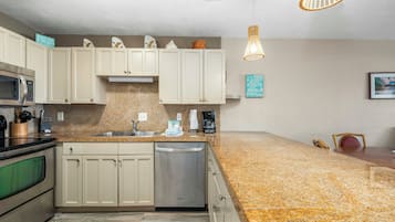 Una cocina con gabinetes blancos, encimeras de granito, lavavajillas de acero inoxidable y un microondas. Hay dos lámparas colgantes y una cafetera en la encimera.