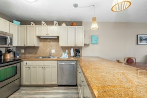 A kitchen with white cabinets, granite countertops, a stainless steel dishwasher, and a microwave. There are two hanging pendant lights and a coffee maker on the counter. - Banyan Harbor Resort (Lihue)
