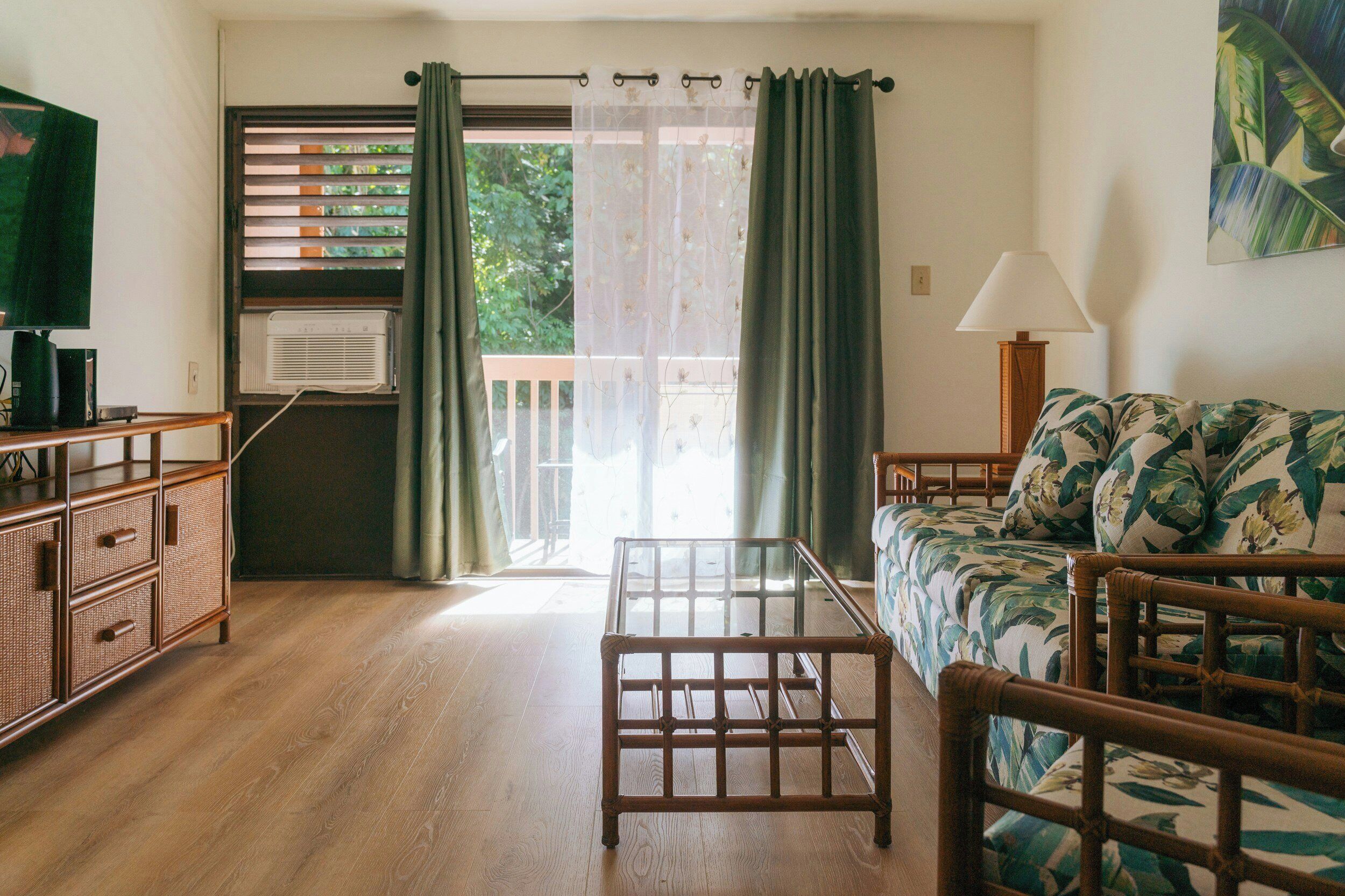A room with a wooden floor, a sofa with floral upholstery, a wooden chair, a small table, a television, an air conditioning unit, and a window with a view of greenery.
